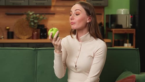 Young Brunette Woman Eats a Green Apple Sitting on a Green Sofa in the Living Room