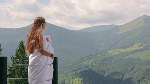 Jovem mulher enrolada em cobertor aprecia a vista da montanha em pé na varanda com vista para os picos dos Cárpatos