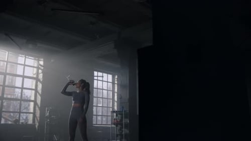 Powerful sportswoman hydrating after intense cardio workout in dark fitness gym