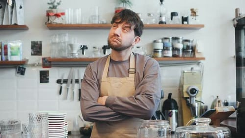 A Barista Stands at the Counter in a Cafe with Arms Crossed Colleagues Move Around Preparing Drinks