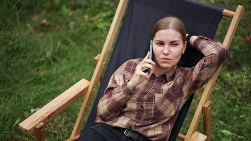 Relaxed happy Caucasian lady speaks on the phone lying in the garden chair.