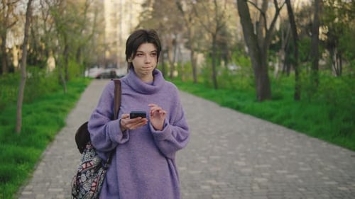 Young Woman Walks on a Pathway While Using a Smartphone in a Green Park Setting During Daylight