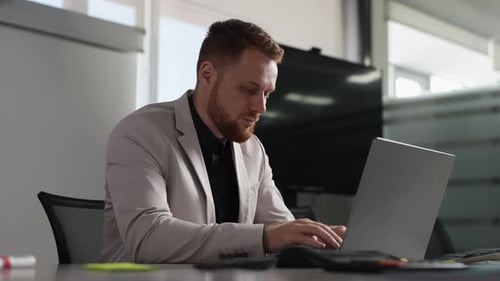 Serious Bearded Businessman in Suit Working on Laptop Computer Sitting at Desk in Modern Office Room