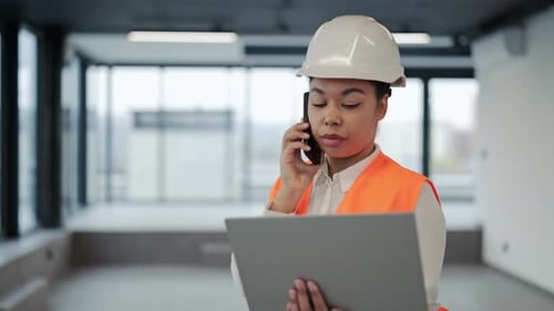Confident Female Architect in Hardhat Reviews Blueprints on Laptop During Phone Call at Construction