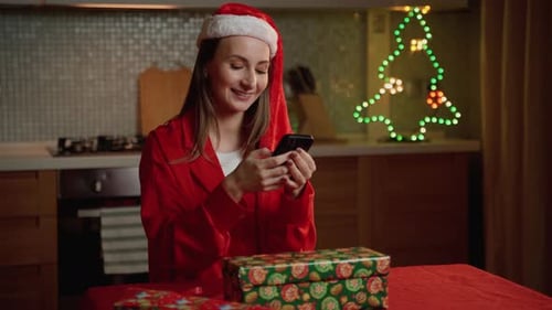 Smiling Young Woman Using Smartphone Next to Wrapped Christmas Gift Indoors