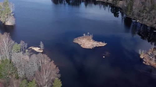 Drone shot orbiting over small island in lake in Sweden during day. Reflections of sky in the lake.