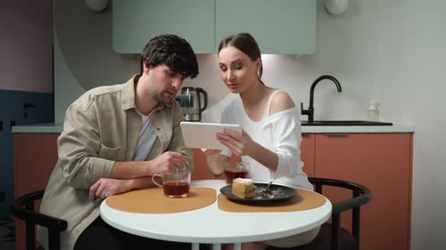 Smiling Young Married Couple is Sitting at a Table in a Home Kitchen Using an Online Store of Modern