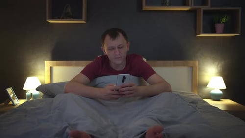 Man Sitting in Bed at Night Uses His Mobile Phone Browses the App Bedroom Interior Front View