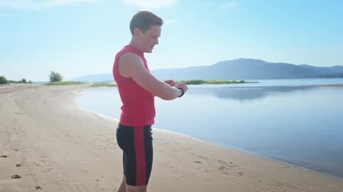 Runner on a beach. Young sportive man prepares for run and sets his watch being a sandy coast of a r