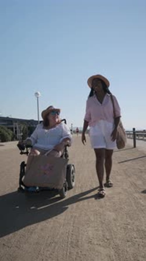 Woman Pushing Friend in Wheelchair on Beach Path in Slow Motion