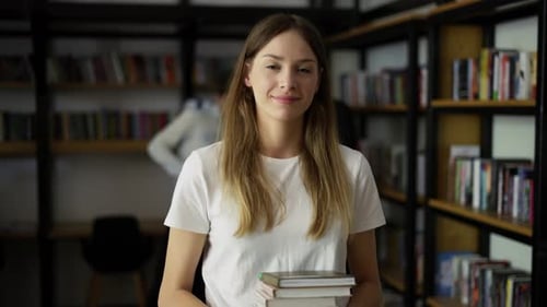 Student Girl Walks Through Library with Stack of Books in Hands Shelves with Book Front View
