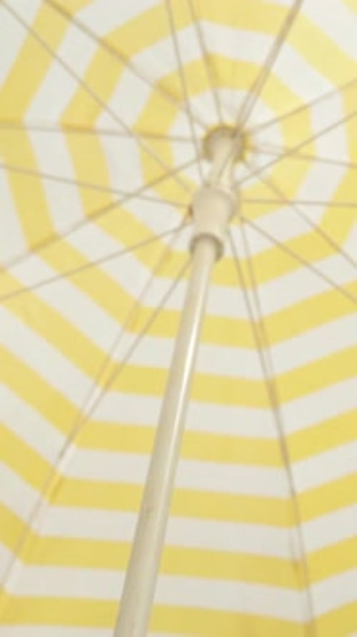 Vertical video. A beach umbrella with white and yellow stripes provides shade from the sun.