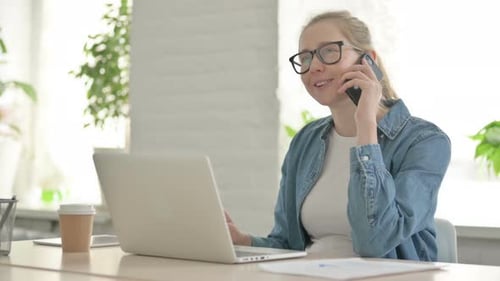 Beautiful Young Woman Talking on Phone While Using Laptop in Office