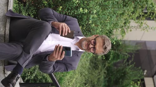 Man Sits With Phone and Coffee Outdoors