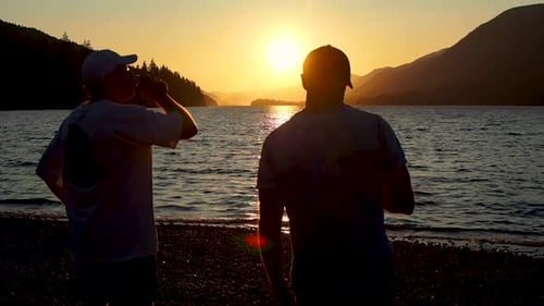 two guys on beach drinking watching the sunset with lake and mountains in the background