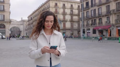 Cheerful Curly Young Woman in Casual Clothes Using Mobile Phone at City Street
