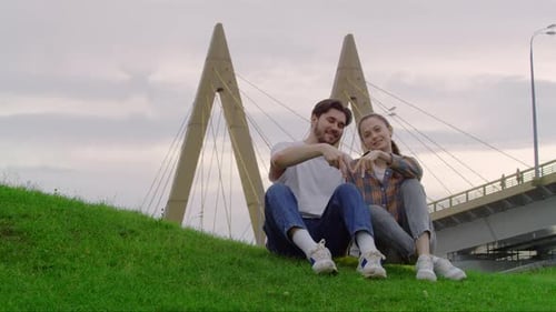 A Young Couple are Sitting in a Park Holding Hands and Looking Straight at the Camera They're Making