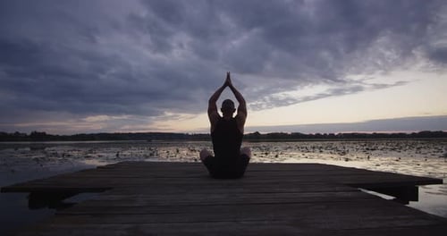 A Young Guy Does Yoga in the Evening in Nature on the River Bank