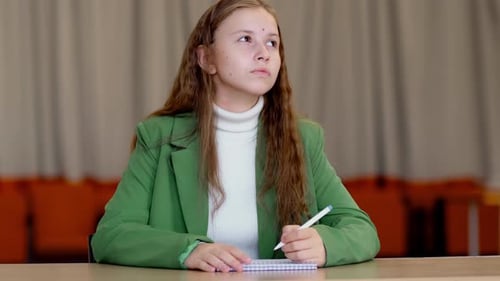 Teenage Girl in Green Blazer Sits at Desk with Notebook and Pen Looks Up to the Side Trying to