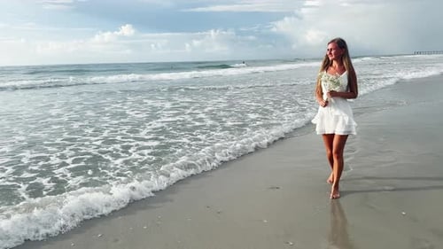Woman in a White Dress Holds Flowers By the Serene Beach