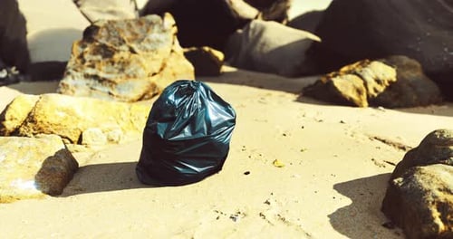Trash Bag Left on a Sandy Beach Surrounded By Rocks During Daylight Hours