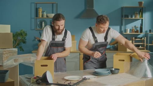 Workers of Moving Company With Uniforms Carefully Packing Dishes at Boxes