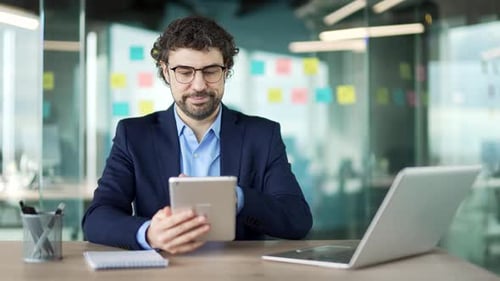 Confident businessman is using digital tablet while sitting at workplace in business office. Man