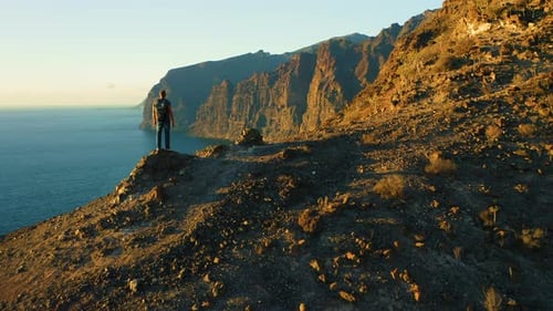 Man Backpacker Stands on Mountain Top Ocean Coast at Evening Sky Sunset Background