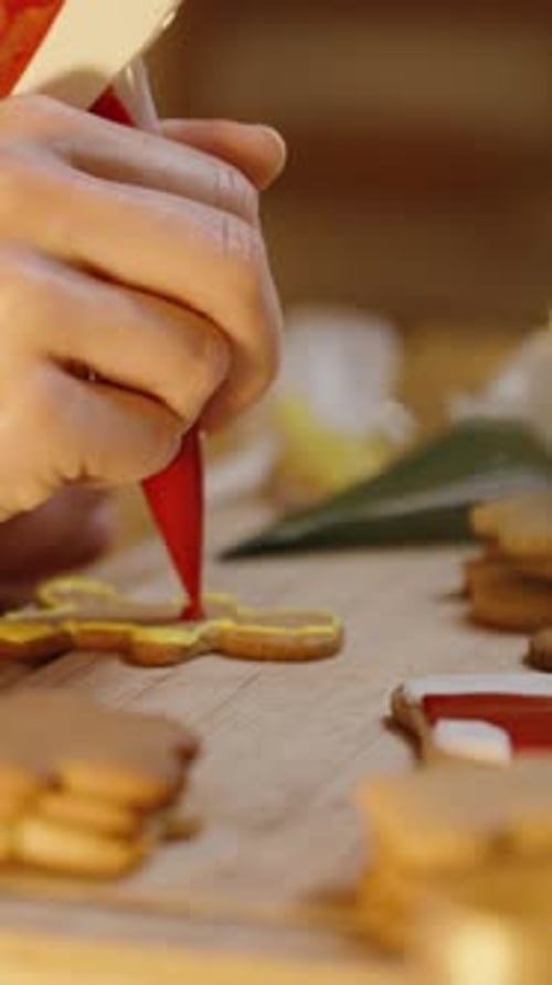 Vertical video. A Woman in a Festive Red Sweater Adds Red Details to a Gingerbread Man Cookie.