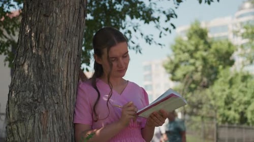 Woman Writing in Notebook Leaning Against Tree