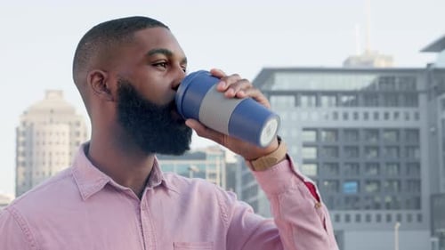 Bearded African American male drinking coffee from travel mug with a background of city buildings