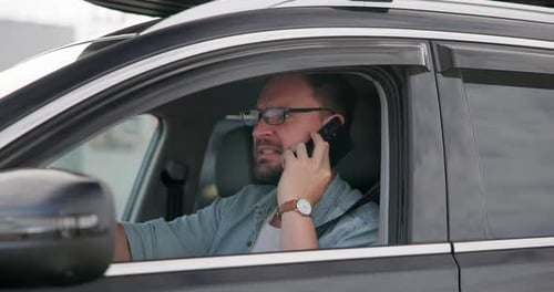 Young Caucasian Man Sits in a Car on a Parking Lot and Talking on the Phone