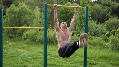 Athletic Man Trains Abdominal Muscles Raising Legs on Horizontal Bar at Sports Ground Young