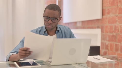 Worried Man Reading Documents at Desk with Laptop