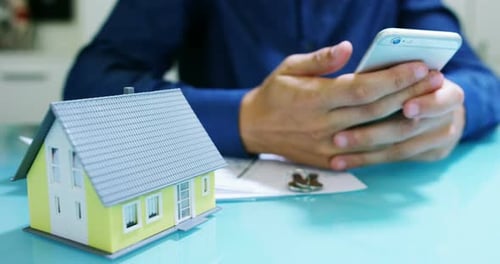 Person Using Smartphone with House Model and Keys