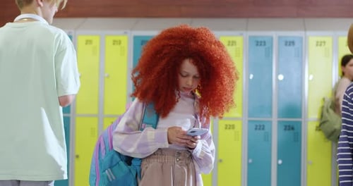 Redhead Girl Using Modern Smartphone in School Hallway While Other Students Walking Around Her