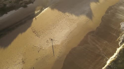Luftaufnahme von oben nach unten eines jungen Surfers, der bei Sonnenuntergang aus dem Wasser kommt und am Sandstrand spaziert