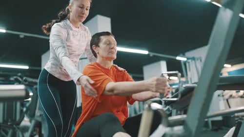 Senior Woman Using Exercise Machine With Trainer Assistance