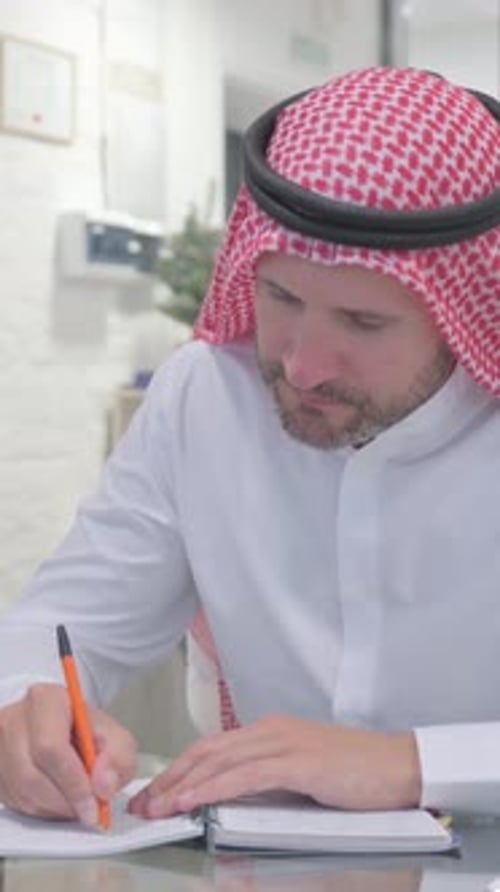 Man Wearing Keffiyeh Writing in Notebook at Workplace