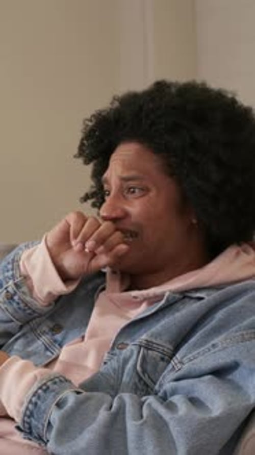 Vertical video: Reacting to worry, adult man pressing knuckle to mouth on couch denim jacket hoodie