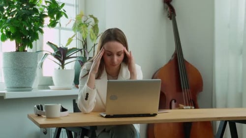 Tired Businesswoman in Stress Works at a Laptop While Sitting at a Table at Home and Holds Her Hand
