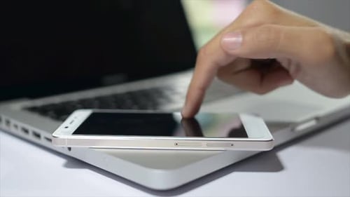Hand Swiping Smartphone Resting on Silver Laptop