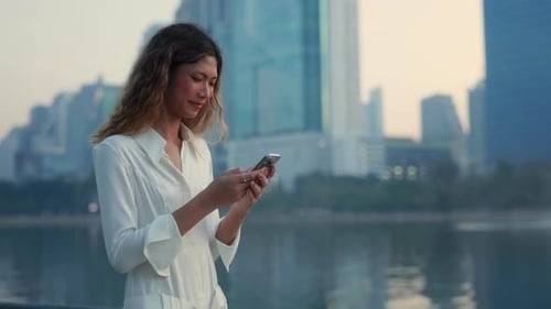 Portrait of Happy Asian Woman Reads Message on Smartphone on City Waterfront