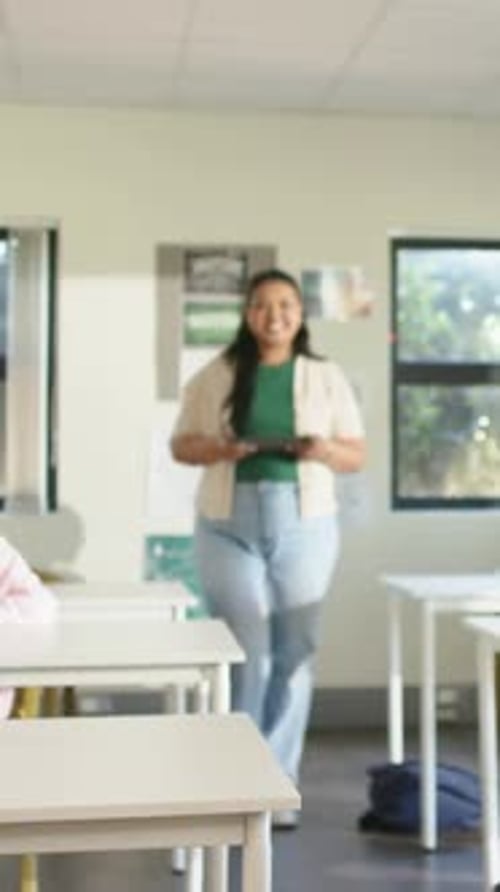 Vertical video: Entering rear, woman in green top walking down aisle in class with tablet to teach