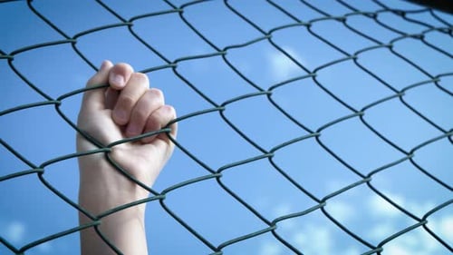 Hand Gripping Chain Link Fence Against Blue Sky