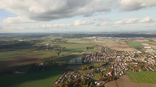 Flying in blue sky below cumulus clouds over green fields and houses on approaching Prague