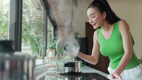Happy Asian woman in green sleeveless open lid from steaming pot checking and smelling food cooking