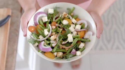 Overhead hands of biracial woman holding bowl of feta salad in kitchen, copy space, slow motion