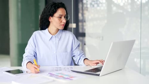 Busy serious young female accountant works using laptop takes notes while sitting at desk