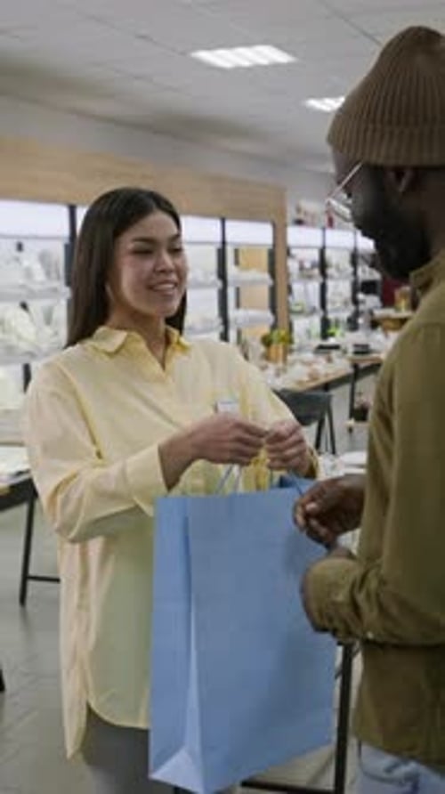 Cheerful Saleswoman Handing Shopping Bag to Black Man in Tableware Store